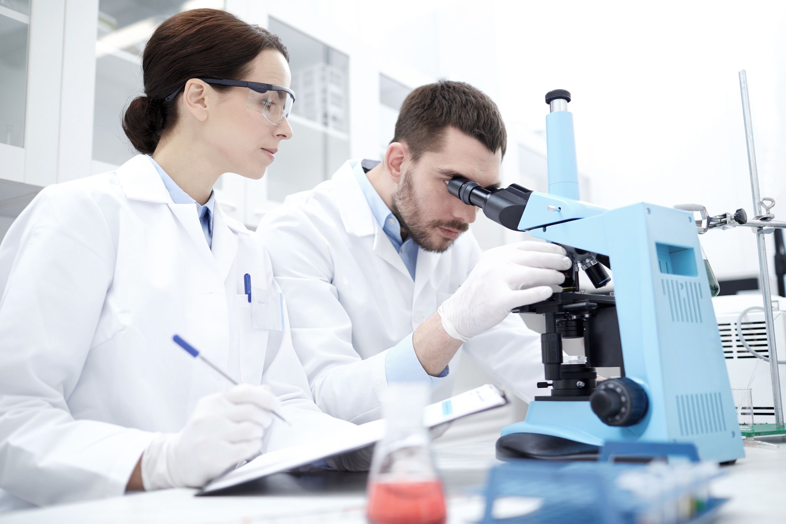 science, chemistry, technology, biology and people concept - young scientists with microscope making test or research in clinical laboratory and taking notes